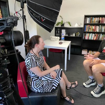 Two people sitting in a room facing eachother, surrounded by filming equipment including a camera and light diffuser 