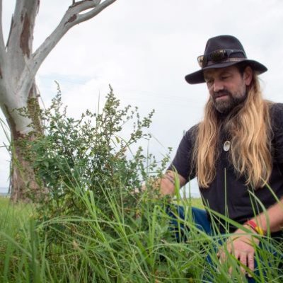 A bearded man with long blond hair wearing a black hat and shirt bends down to look at the long grass