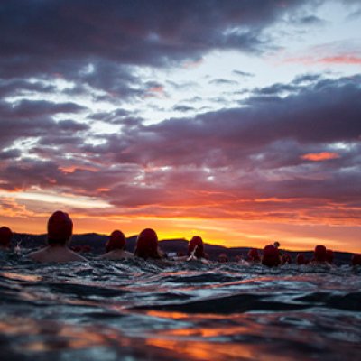 A sunrise photo taken from the water with swimmers in silhouette against a cloudy orange sky.