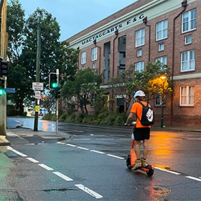 A man in high-vis and wearing a helmet and backpack rides an e-scooter across a Brisbane road.