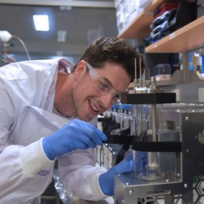 a man in a white lab coat and glasses adjusts a glass cylinder surrounded by stainless steel equipment