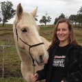 Carissa Horsey, a young woman, standing and smiling next to her light-coloured horse in a field