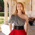 A young woman with long blonde hair standing under a sandstone walkway surrounded by columns