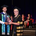 A man and a woman in graduation attire posing on stage - the man holding a cylinder 