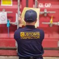 a man in a blue shirt with Customs Inspector on it looks at a red shipping container