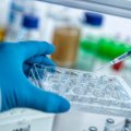 A gloved hand holds a tray of vials as a pipette hovers in a laboratory setting.