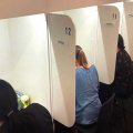 Women sitting behind desks in individual booths with their backs to the camera tasting food