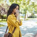 Woman in yellow jacket blowing her nose in a tissue standing in a city park with trees in the background 