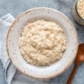 a bowl of porridge sits next to a wooden spoon and a jar of uncooked oats