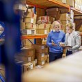 A man and a woman look at a clipboard in a factory while surrounded by boxes