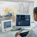 Man in white coat looking at radiology images with patient in CT scanner in background. Adobe.