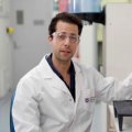 a man in a white lab coat sits next to a multicoloured model