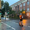 A man in high-vis and wearing a helmet and backpack rides an e-scooter across a Brisbane road.