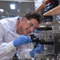 a man in a white lab coat and glasses adjusts a glass cylinder surrounded by stainless steel equipment