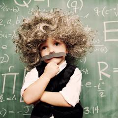 A young child dressed as Einstein stands in front of a chalk board covered in equations
