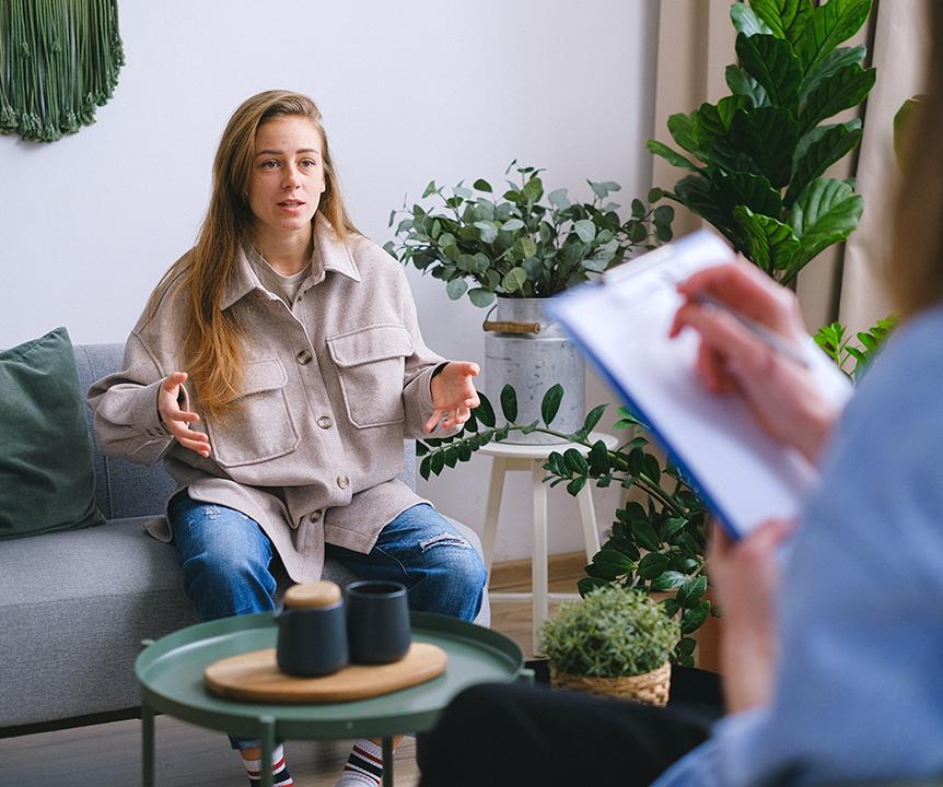 A woman sitting on a couch complaining to another person, who is holding a clipboard in hand and taking notes.