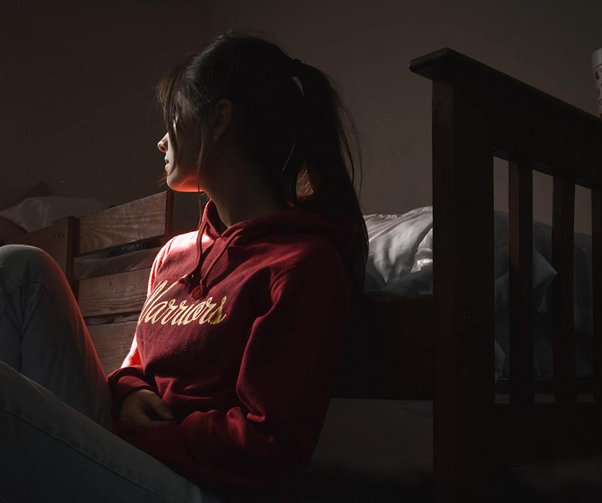 A girl sitting in the dark by her bed with her face turned away from the camera. 