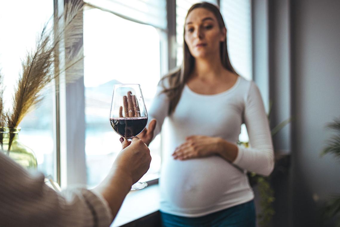 A hand holding out a glass of red wine to a pregnant woman, who is signalling she does not want it