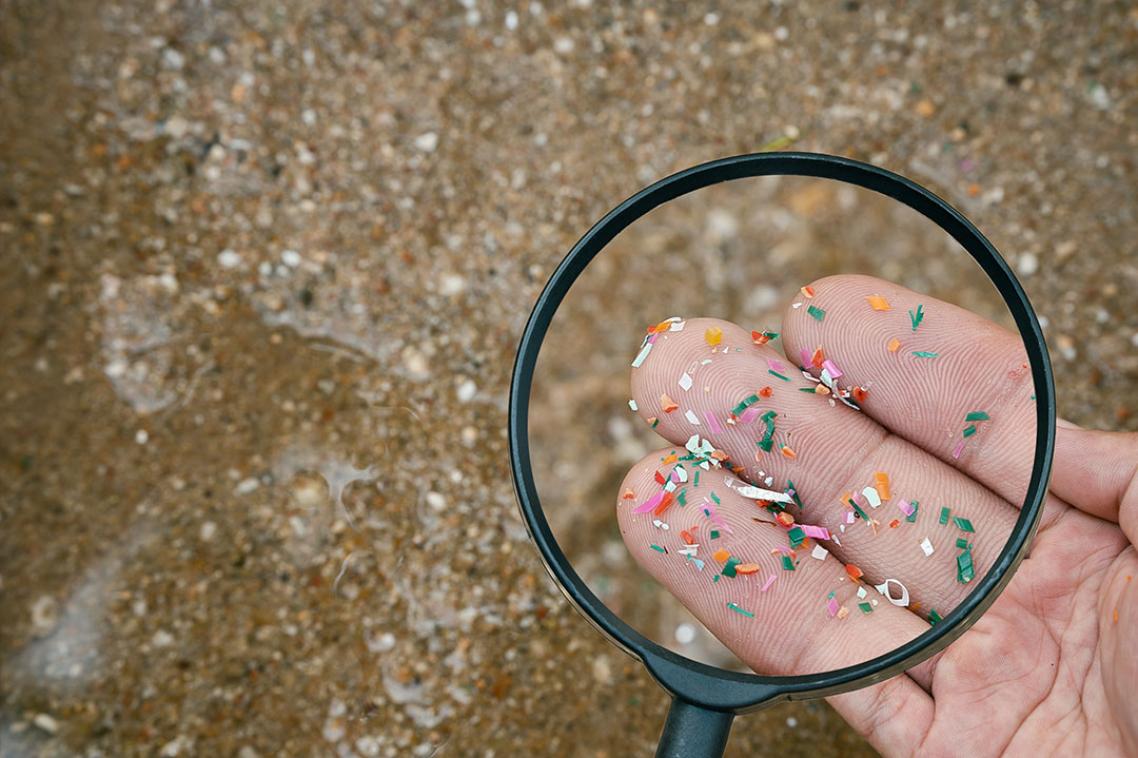A magnifying glass on a hand holding tiny pieces of plastic with a beach in the background