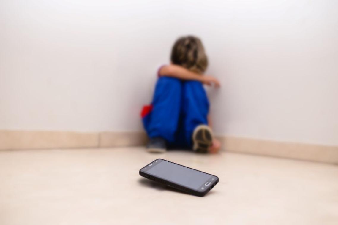 A boy with his head in his knees leaning against a wall, a phone sitting in front of him