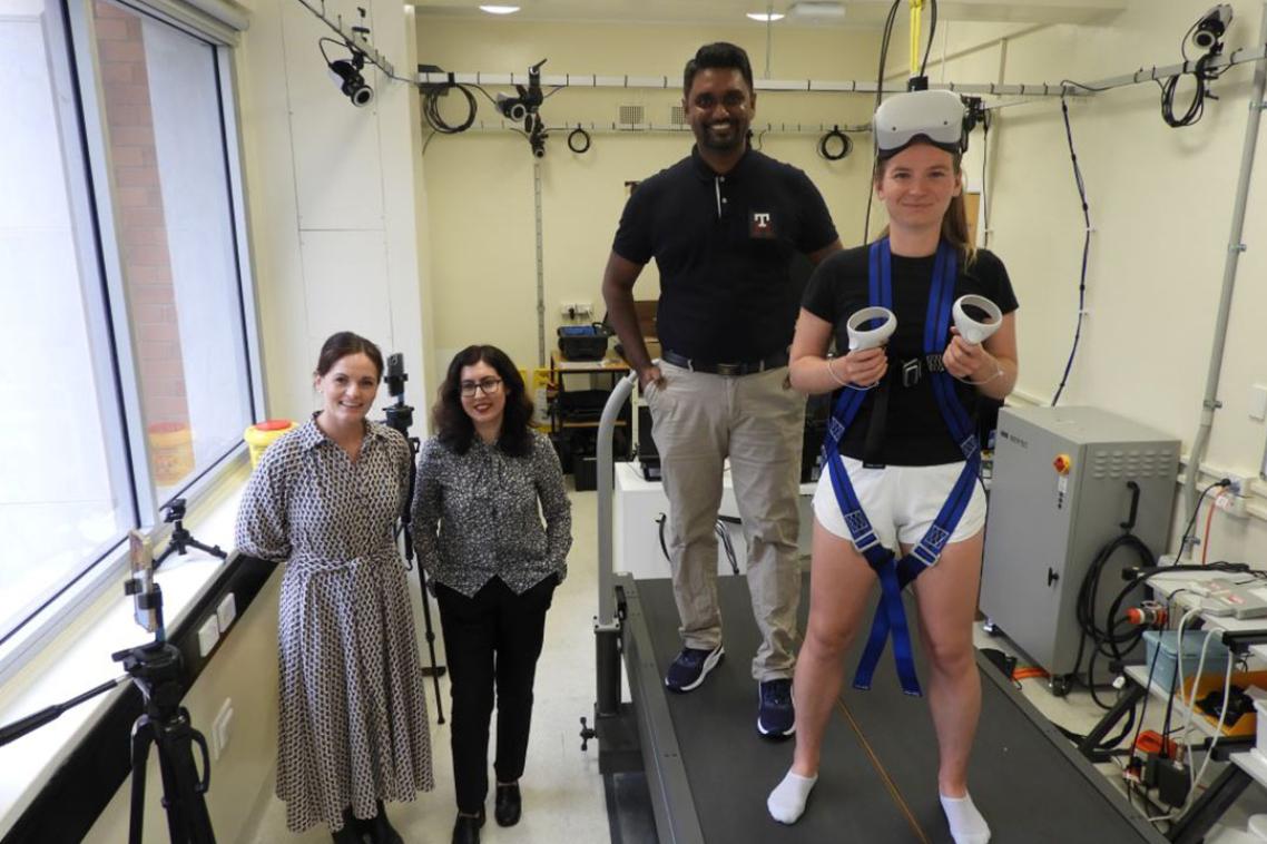 Four people standing in a lab, one in a harness wearing VR goggles and holding two remotes
