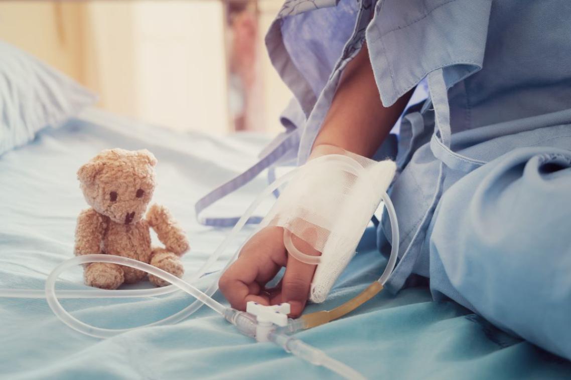 A close up of a child's hand on a hospital bed, attached to tubing, a small teddy bear placed next to it